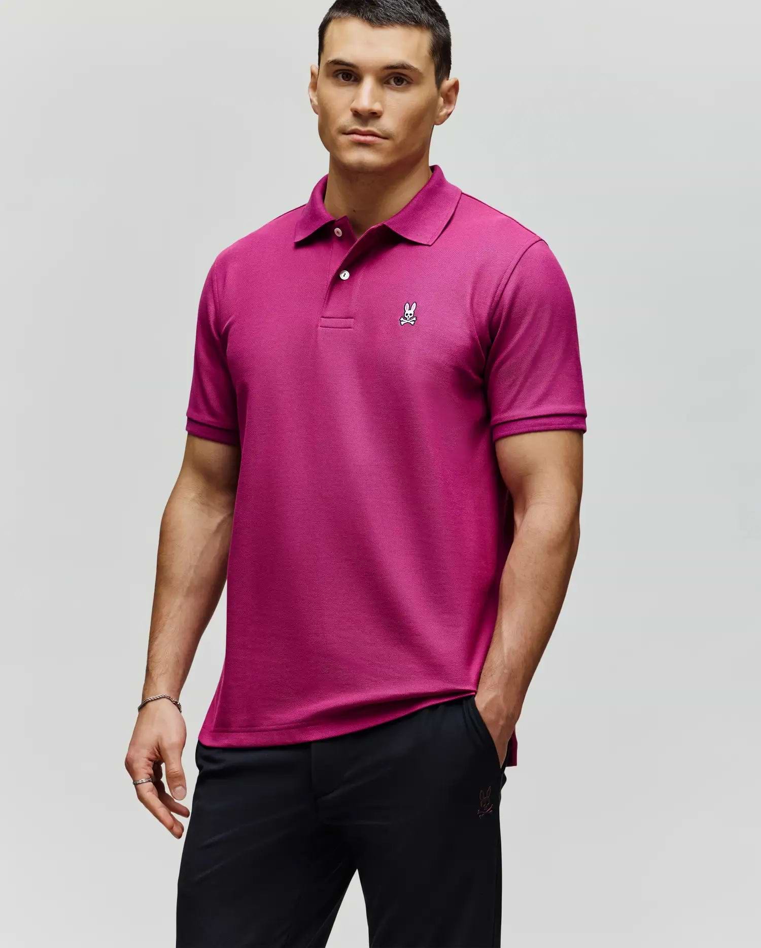 A man wears a Psycho Bunny MENS CLASSIC PIQUE POLO - B6K001F200 in bright magenta, featuring an embroidered Bunny logo on the chest, paired with black pants, against a plain light background.