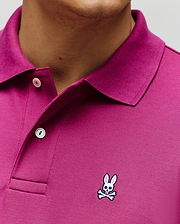 A close-up of a person wearing the Psycho Bunny MENS CLASSIC PIQUE POLO (B6K001F200) in magenta, made from soft Pima cotton, with white buttons and the embroidered Bunny logo with crossbones on the chest.