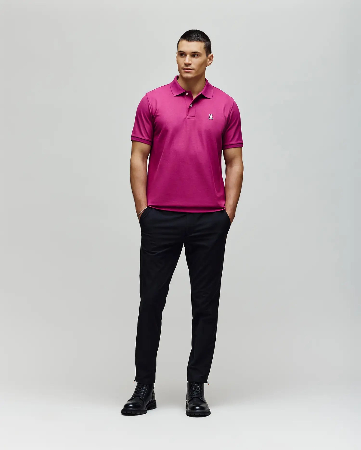 A man stands against a plain background wearing Psycho Bunny’s MENS CLASSIC PIQUE POLO - B6K001F200 in bright pink, featuring the embroidered Bunny logo, with black pants and shoes, hands in pockets and looking to the side.