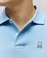 Light Blue Mens Classic Pique Polo with a textured pique knit, featuring a two-button placket and a small, embroidered white and pink Psycho Bunny skull logo on the left chest.