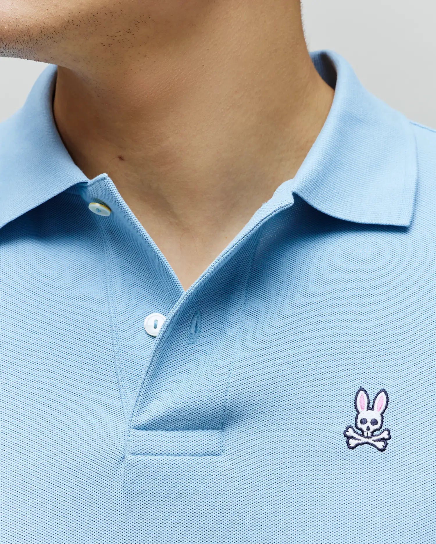 Light Blue Mens Classic Pique Polo with a textured pique knit, featuring a two-button placket and a small, embroidered white and pink Psycho Bunny skull logo on the left chest.