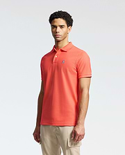 A person stands wearing the Psycho Bunny MENS CLASSIC PIQUE POLO - B6K001F200 in bright orange with an embroidered bunny logo on the chest, paired with light beige pants against a plain, light-colored background.