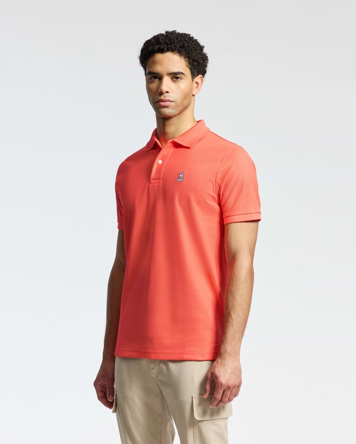 A person stands wearing the Psycho Bunny MENS CLASSIC PIQUE POLO - B6K001F200 in bright orange with an embroidered bunny logo on the chest, paired with light beige pants against a plain, light-colored background.
