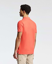 A man with short curly hair wears a Psycho Bunny coral MENS CLASSIC PIQUE POLO - B6K001F200, featuring a subtle bunny logo. He pairs it with beige pants and gazes left against a plain white background.