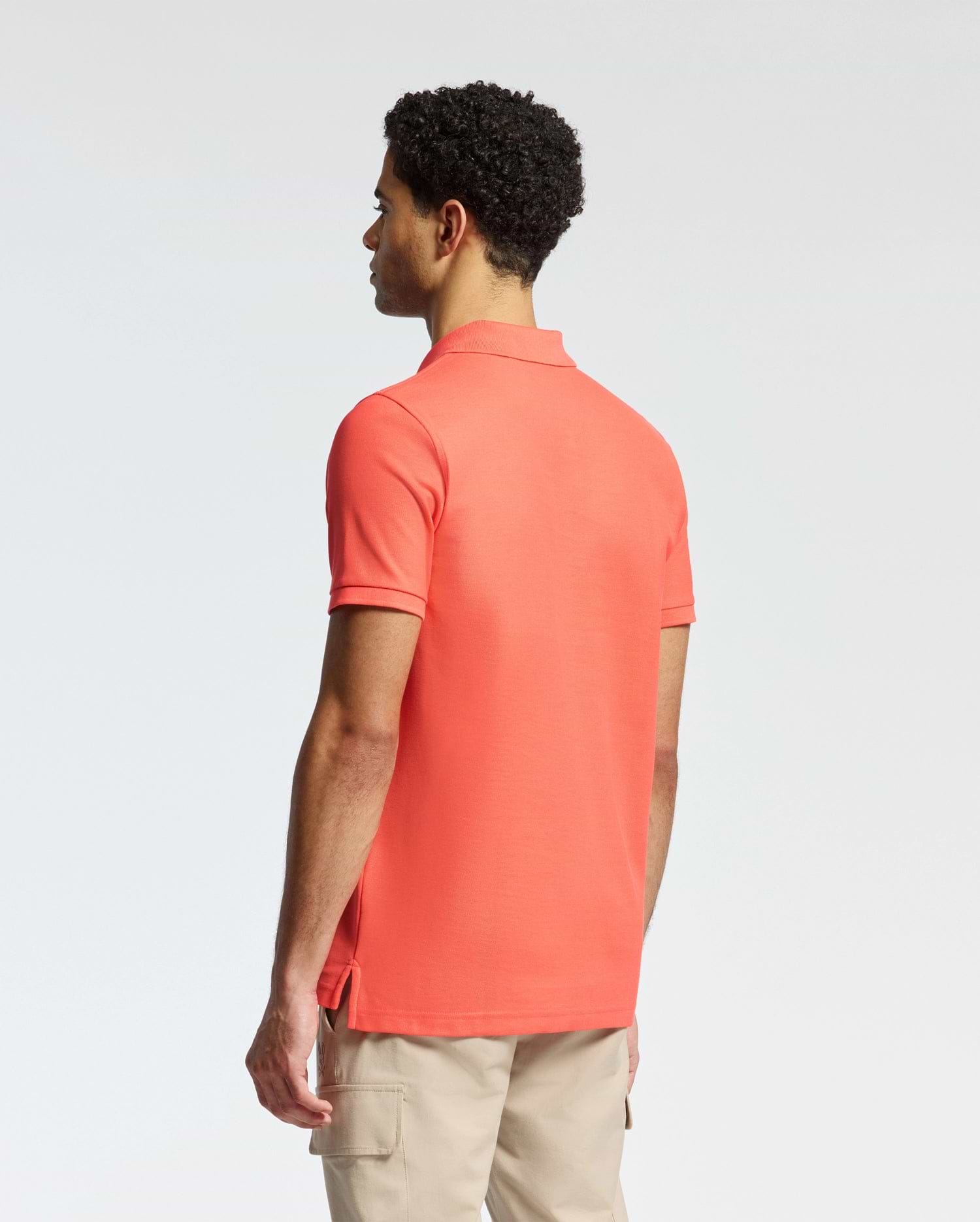 A man with short curly hair wears a Psycho Bunny coral MENS CLASSIC PIQUE POLO - B6K001F200, featuring a subtle bunny logo. He pairs it with beige pants and gazes left against a plain white background.