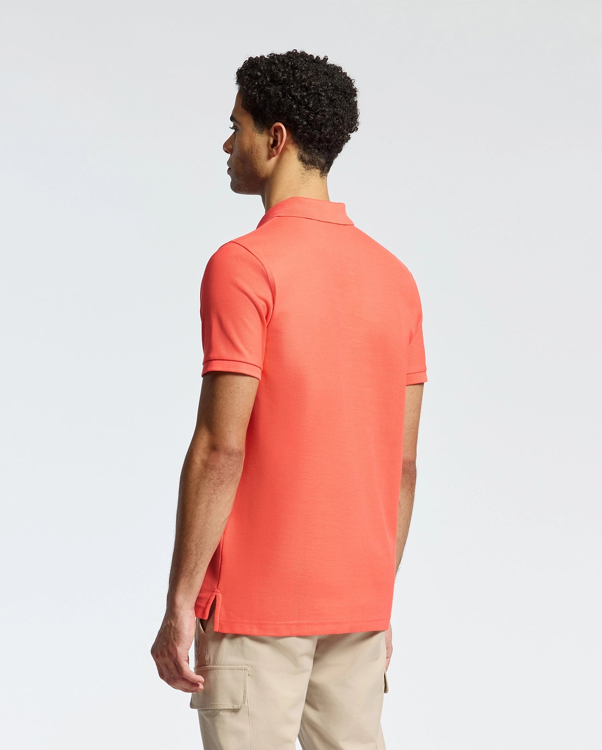 A man with short curly hair wears a Psycho Bunny coral MENS CLASSIC PIQUE POLO - B6K001F200, featuring a subtle bunny logo. He pairs it with beige pants and gazes left against a plain white background.