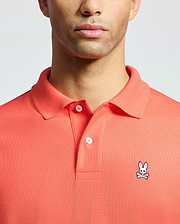 A man models the Psycho Bunny MENS CLASSIC PIQUE POLO - B6K001F200 in coral, made from soft Pima cotton. The upper torso shot showcases the small embroidered bunny with crossbones on the left chest.