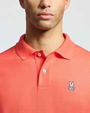 A man models the Psycho Bunny MENS CLASSIC PIQUE POLO - B6K001F200 in coral, made from soft Pima cotton. The upper torso shot showcases the small embroidered bunny with crossbones on the left chest.