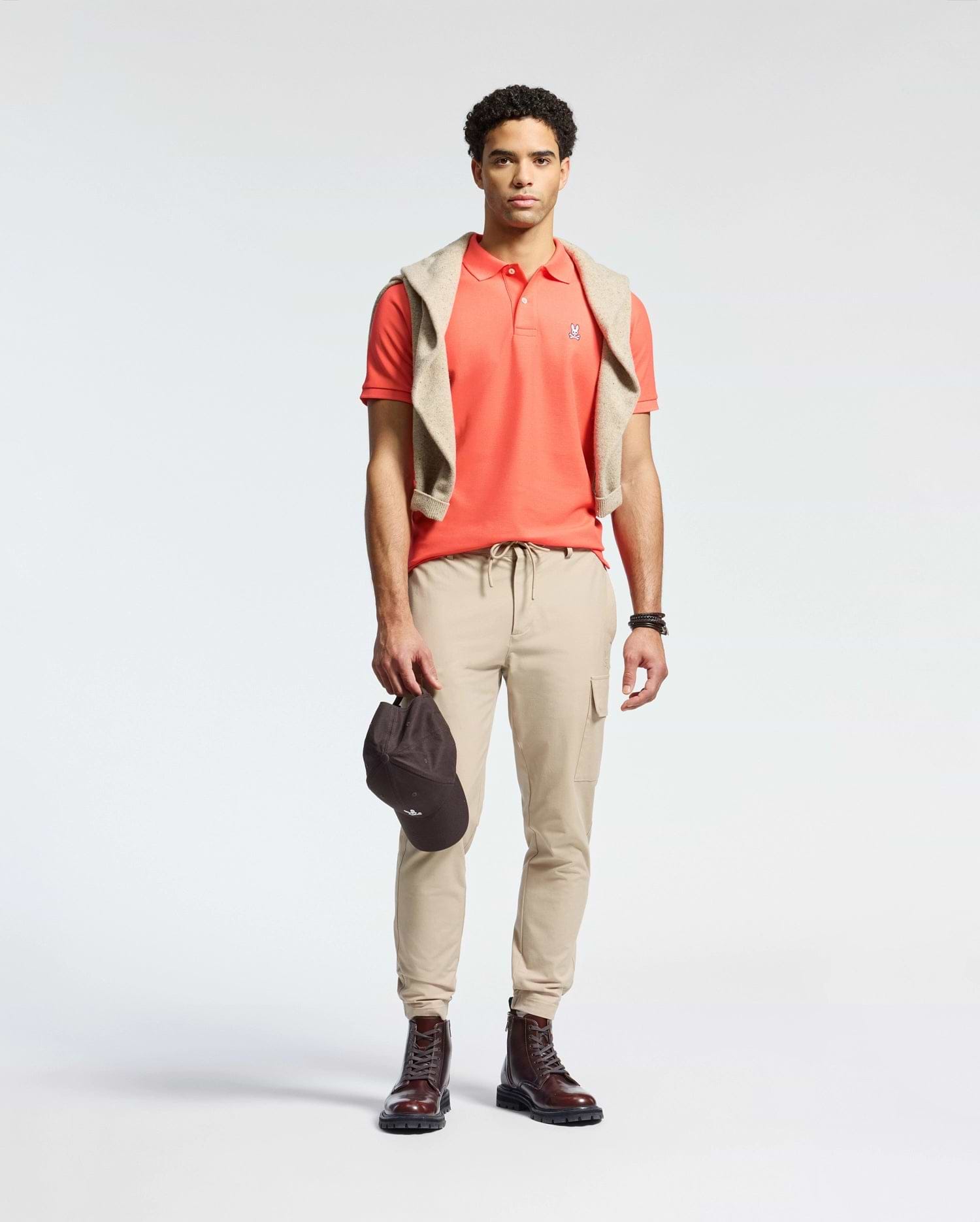 A man in a Psycho Bunny MENS CLASSIC PIQUE POLO - B6K001F200 with an embroidered Bunny logo, beige cargo joggers, and brown boots stands against a plain background. A beige Pima cotton sweater is draped over his shoulders as he holds a dark cap in his right hand.