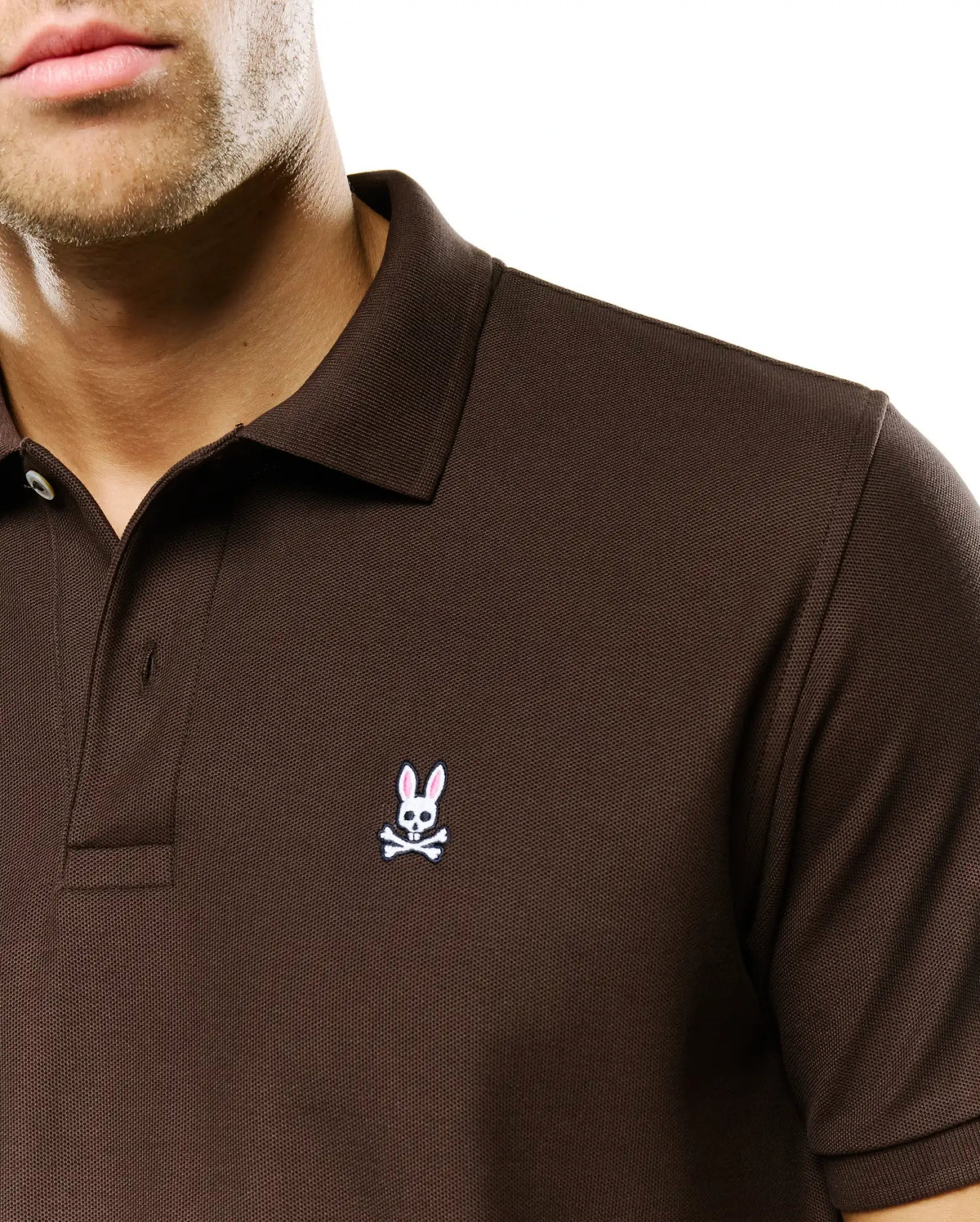 Men's Classic Pima Pique Polo in rich brown, showcasing a classic fit and visible pique texture. A small, embroidered white Psycho Bunny skull with pink ears and black crossbones is on the left chest. (3)