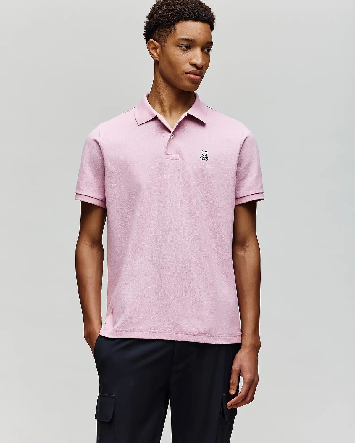 A young man wears the Psycho Bunny MENS CLASSIC PIQUE POLO SHIRT (B6K001F200) in light pink, featuring an embroidered Bunny logo on the chest, paired with dark pants against a plain light gray background.