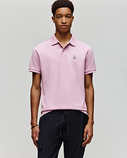 A young man wears the Psycho Bunny MENS CLASSIC PIQUE POLO SHIRT (B6K001F200) in light pink, featuring a small embroidered Bunny logo on the chest, standing against a plain light gray background with his hands by his sides.