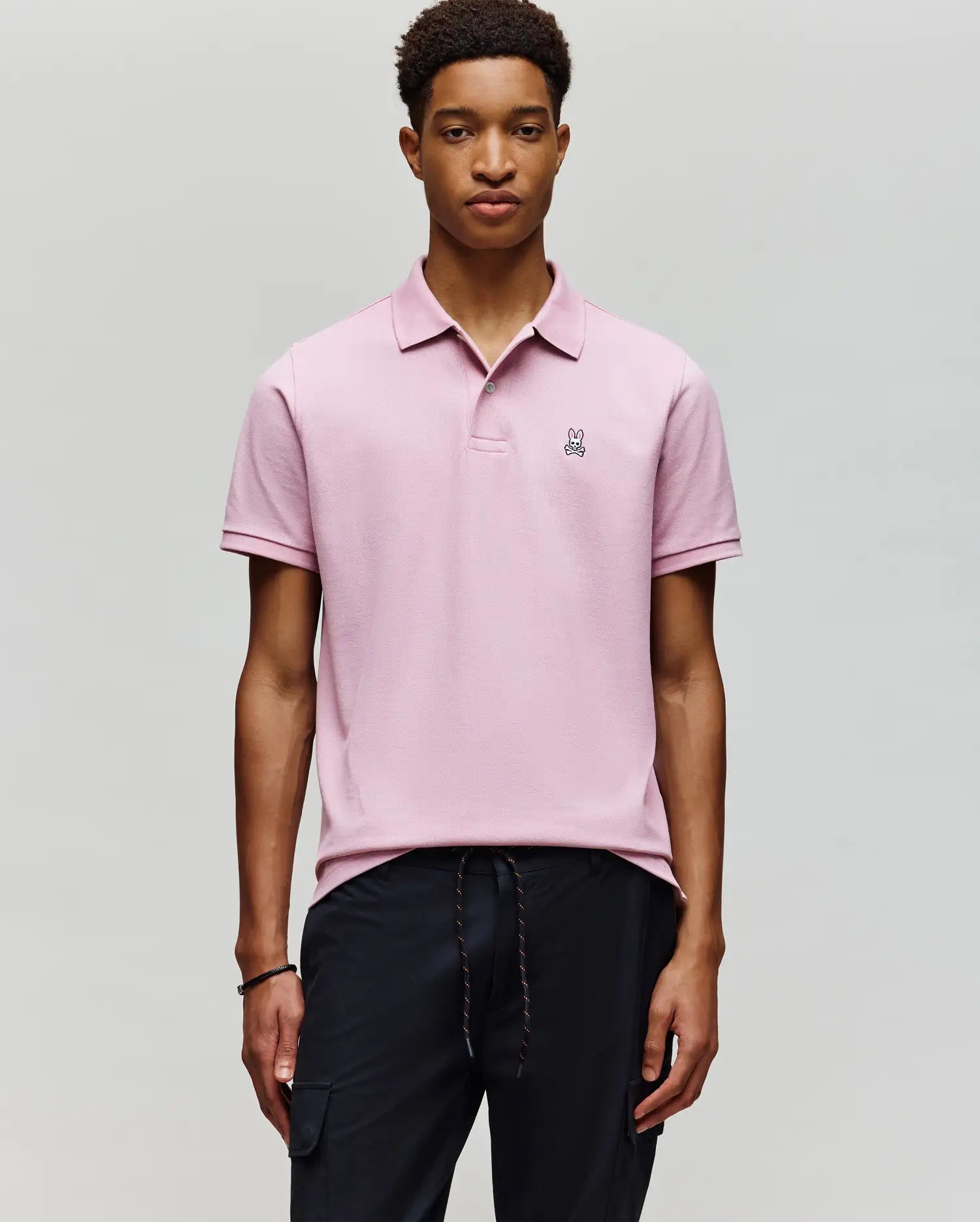 A young man wears the Psycho Bunny MENS CLASSIC PIQUE POLO SHIRT (B6K001F200) in light pink, featuring a small embroidered Bunny logo on the chest, standing against a plain light gray background with his hands by his sides.
