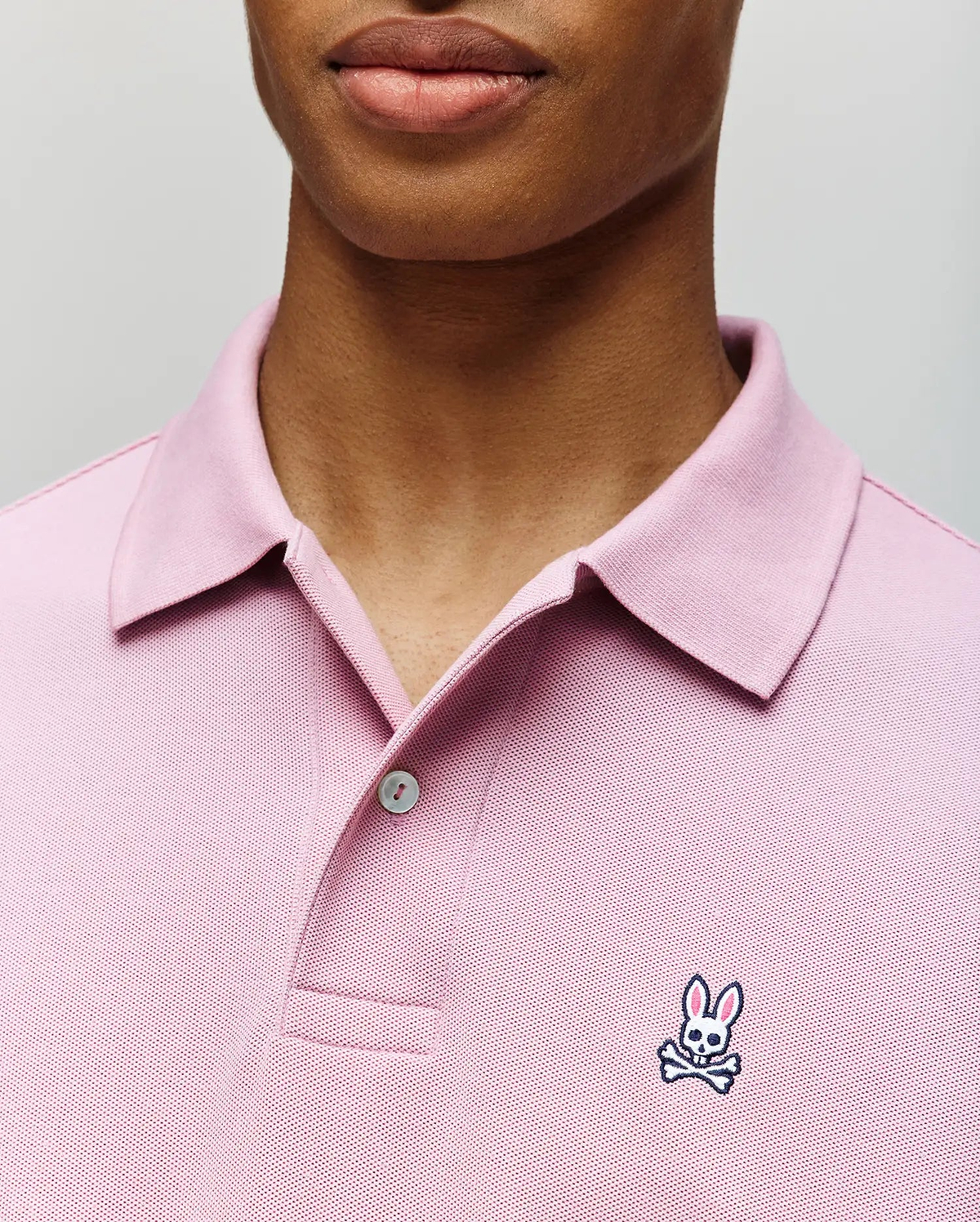 A person wears the Psycho Bunny MENS CLASSIC PIQUE POLO SHIRT (B6K001F200), a light pink men’s polo with a collar, buttons, and the signature embroidered Bunny logo with crossbones on the chest.