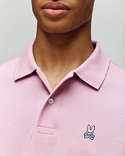 A person wears the Psycho Bunny MENS CLASSIC PIQUE POLO SHIRT (B6K001F200), a light pink men’s polo with a collar, buttons, and the signature embroidered Bunny logo with crossbones on the chest.