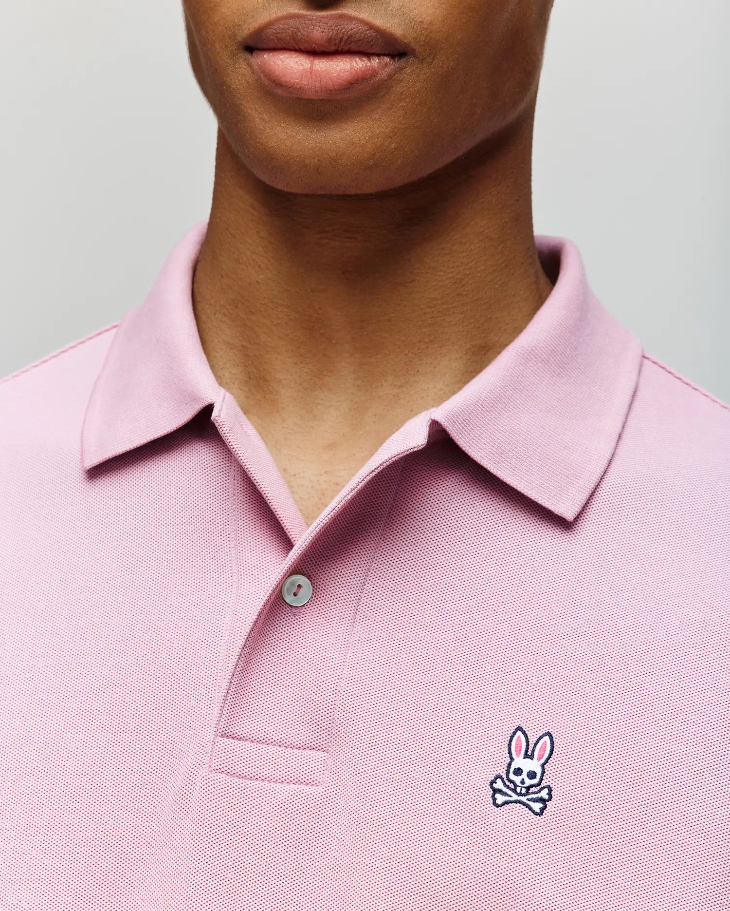 A person wears the Psycho Bunny MENS CLASSIC PIQUE POLO SHIRT (B6K001F200), a light pink men’s polo with a collar, buttons, and the signature embroidered Bunny logo with crossbones on the chest.