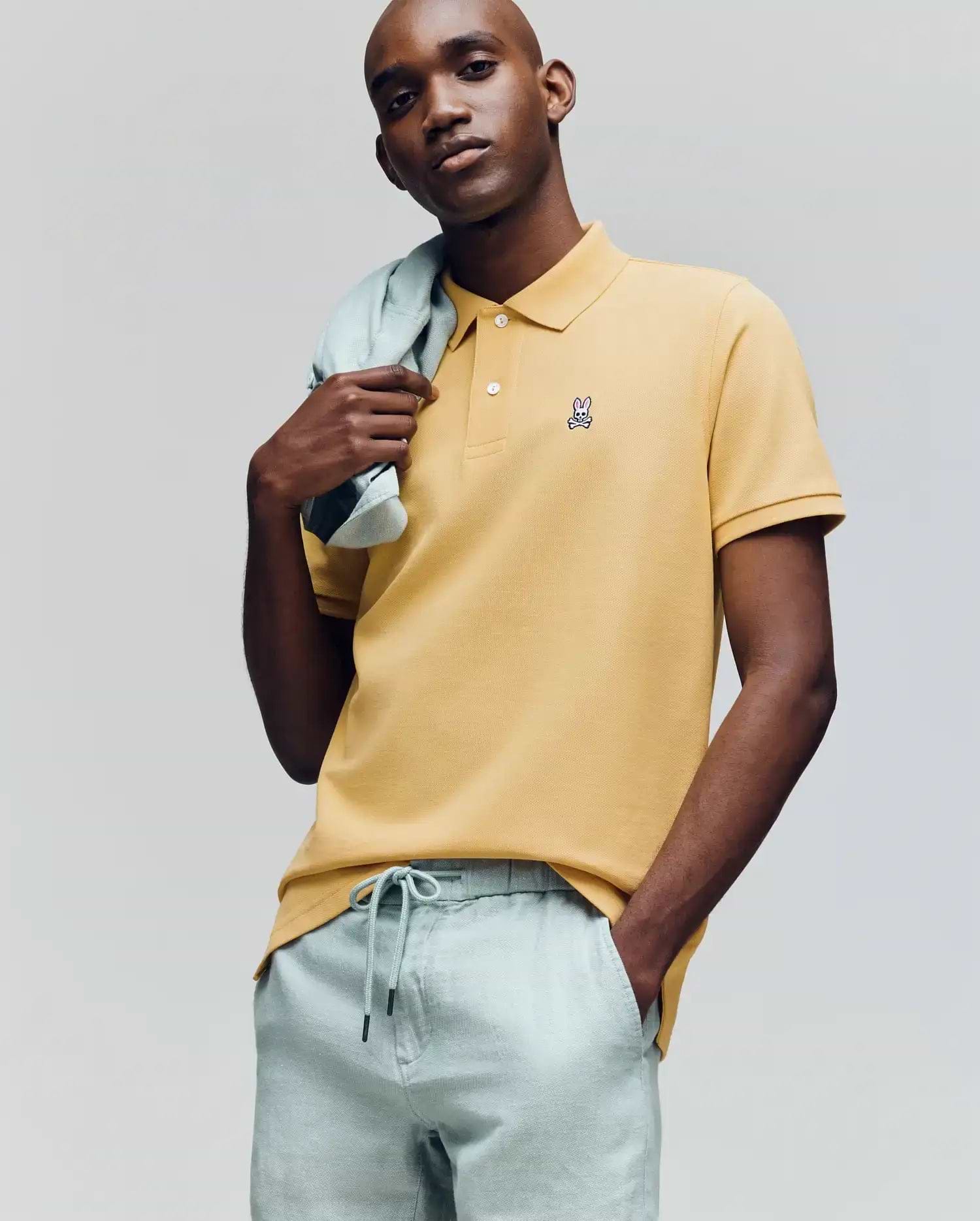 A man in a Psycho Bunny MENS CLASSIC PIQUE POLO (B6K001F200), yellow with an embroidered Bunny logo, and light gray drawstring pants poses with a sweater over his shoulder against a plain background. (2)
