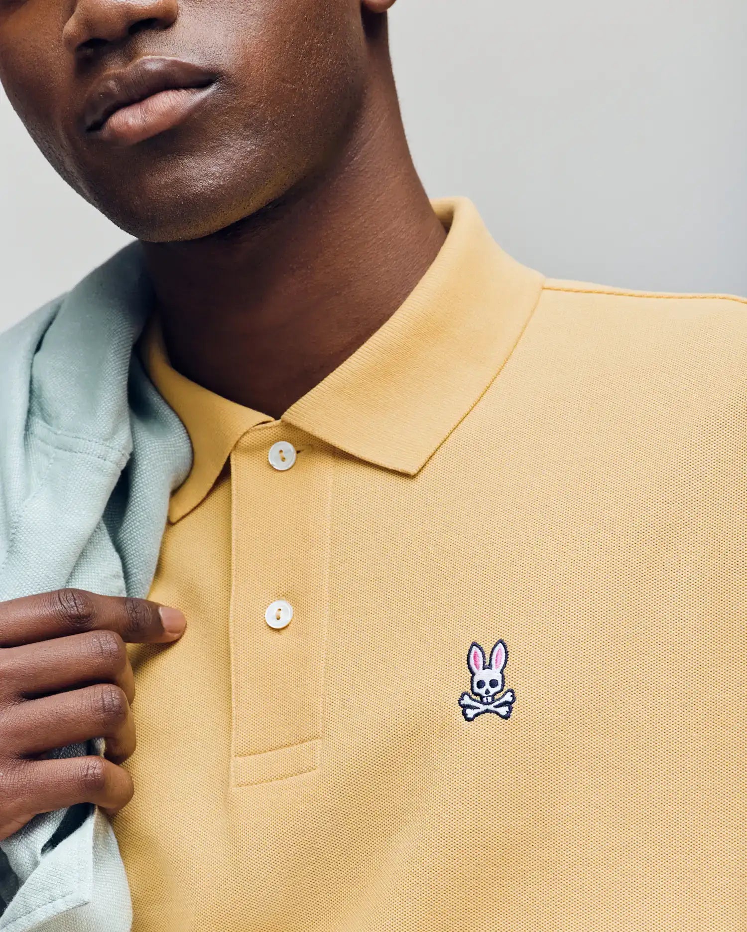 Wearing Psycho Bunny's MENS CLASSIC PIQUE POLO - B6K001F200 in yellow, made from Pima cotton and featuring the embroidered Bunny logo, a person holds a light jacket over their shoulder with only the lower half of their face visible. (3)