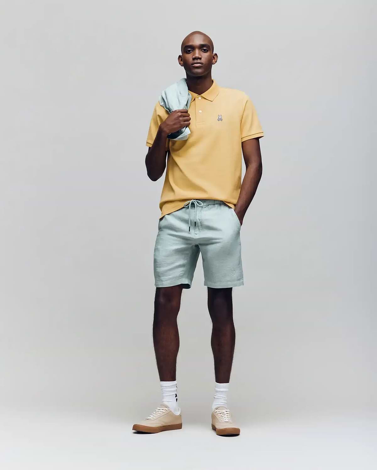 A man stands against a plain background wearing a yellow Psycho Bunny MENS CLASSIC PIQUE POLO - B6K001F200 with an embroidered Bunny logo, light green shorts and matching jacket, plus beige sneakers with white socks. (4)