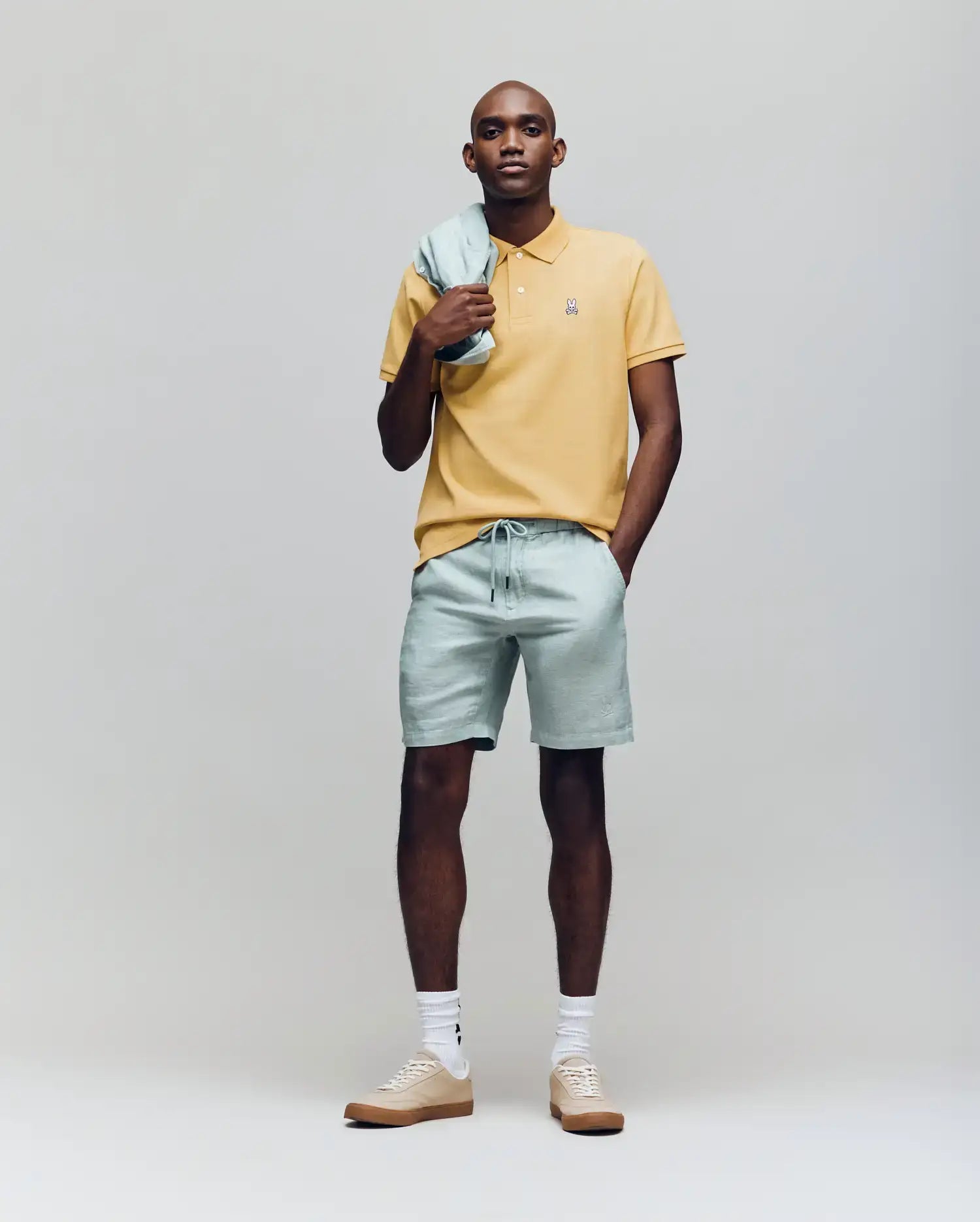 A man stands against a plain background wearing a yellow Psycho Bunny MENS CLASSIC PIQUE POLO - B6K001F200 with an embroidered Bunny logo, light green shorts and matching jacket, plus beige sneakers with white socks. (4)