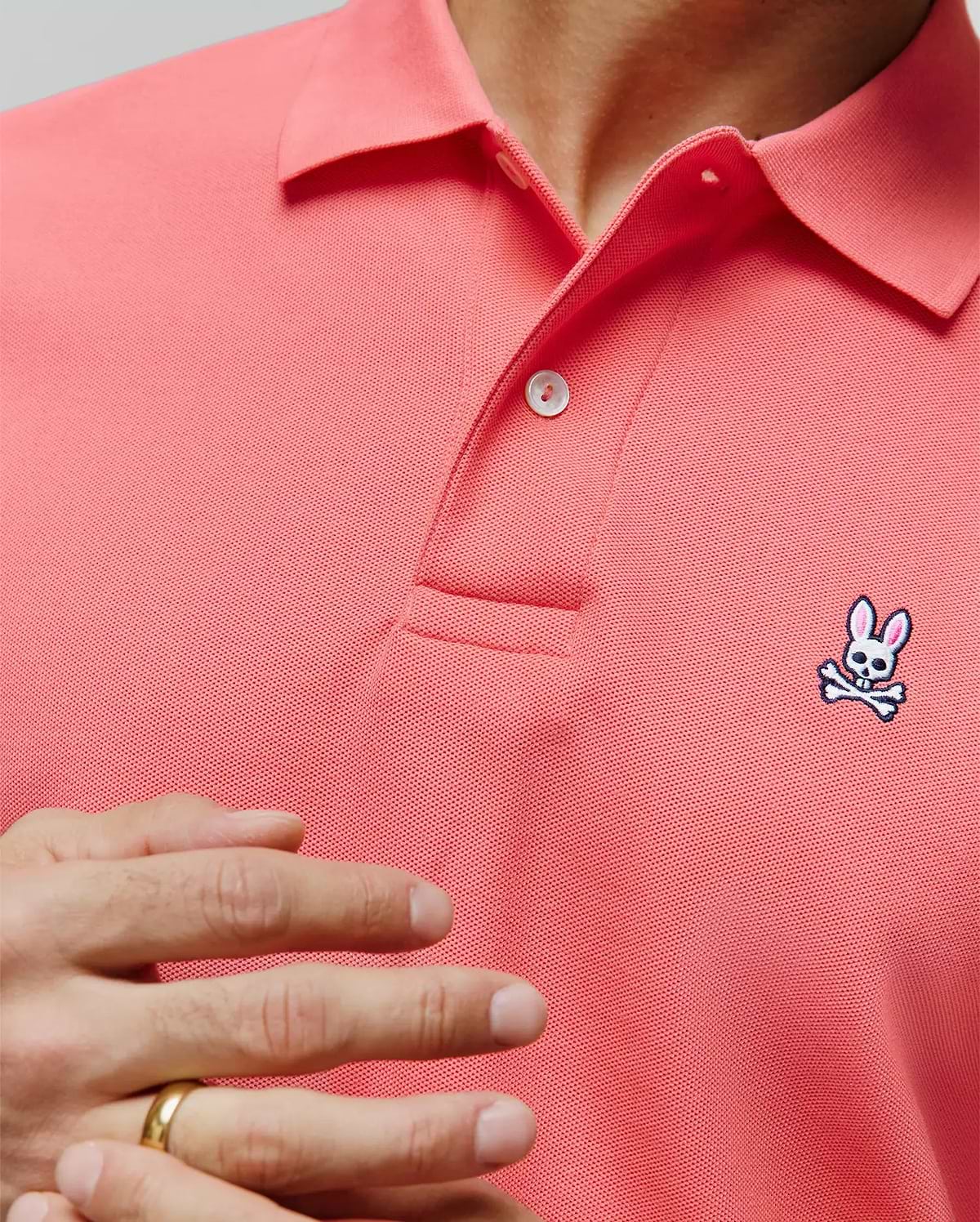 Mens Classic Pique Polo in vibrant coral, featuring a textured pique knit, two white buttons, and a small embroidered Psycho Bunny skull logo on the left chest. (4)