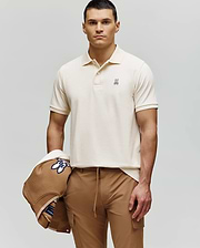 A man with short dark hair wears a Psycho Bunny MENS CLASSIC PIQUE POLO SHIRT (B6K001F200) in cream and tan pants, holding a tan jacket with blue and white design over his left arm, standing against a plain light background.