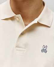 Close-up of a person wearing the Psycho Bunny MENS CLASSIC PIQUE POLO SHIRT - B6K001F200 in cream, featuring two buttons undone and an embroidered Bunny logo with pink ears and crossbones on the left chest.