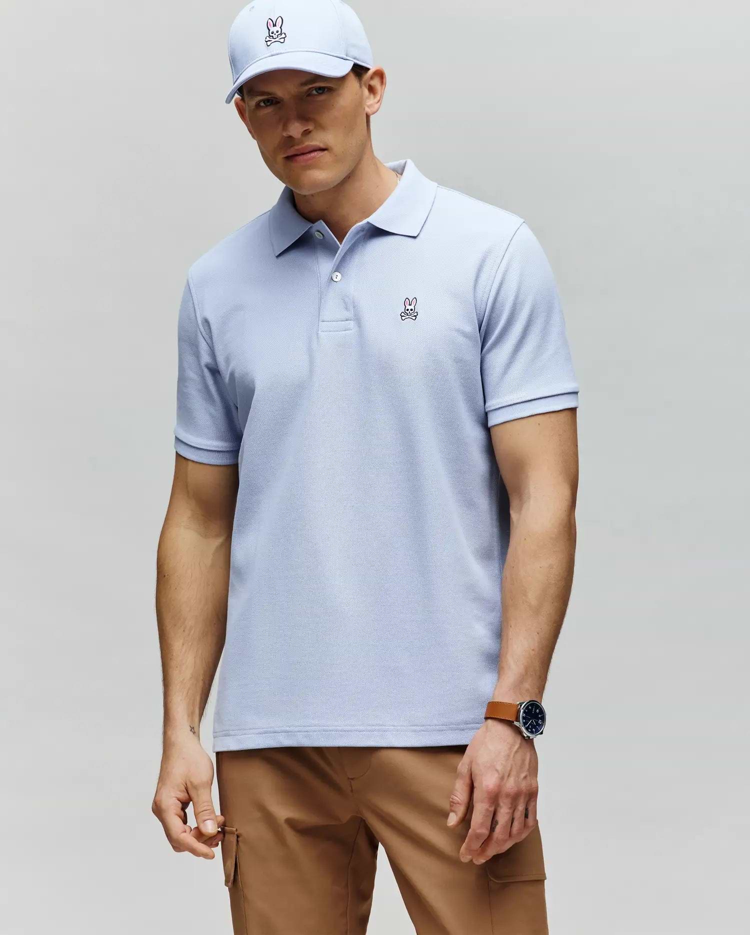 A man stands with his hands by his sides, wearing a Psycho Bunny MENS CLASSIC PIQUE POLO - B6K001F200 in light blue, paired with a matching bunny logo cap, tan pants, and a watch while gazing slightly toward the camera.