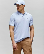 A man in a Psycho Bunny MENS CLASSIC PIQUE POLO - B6K001F200 in light blue with a small bunny logo, paired with a matching cap and tan cargo pants, stands against a gray background looking to the side.