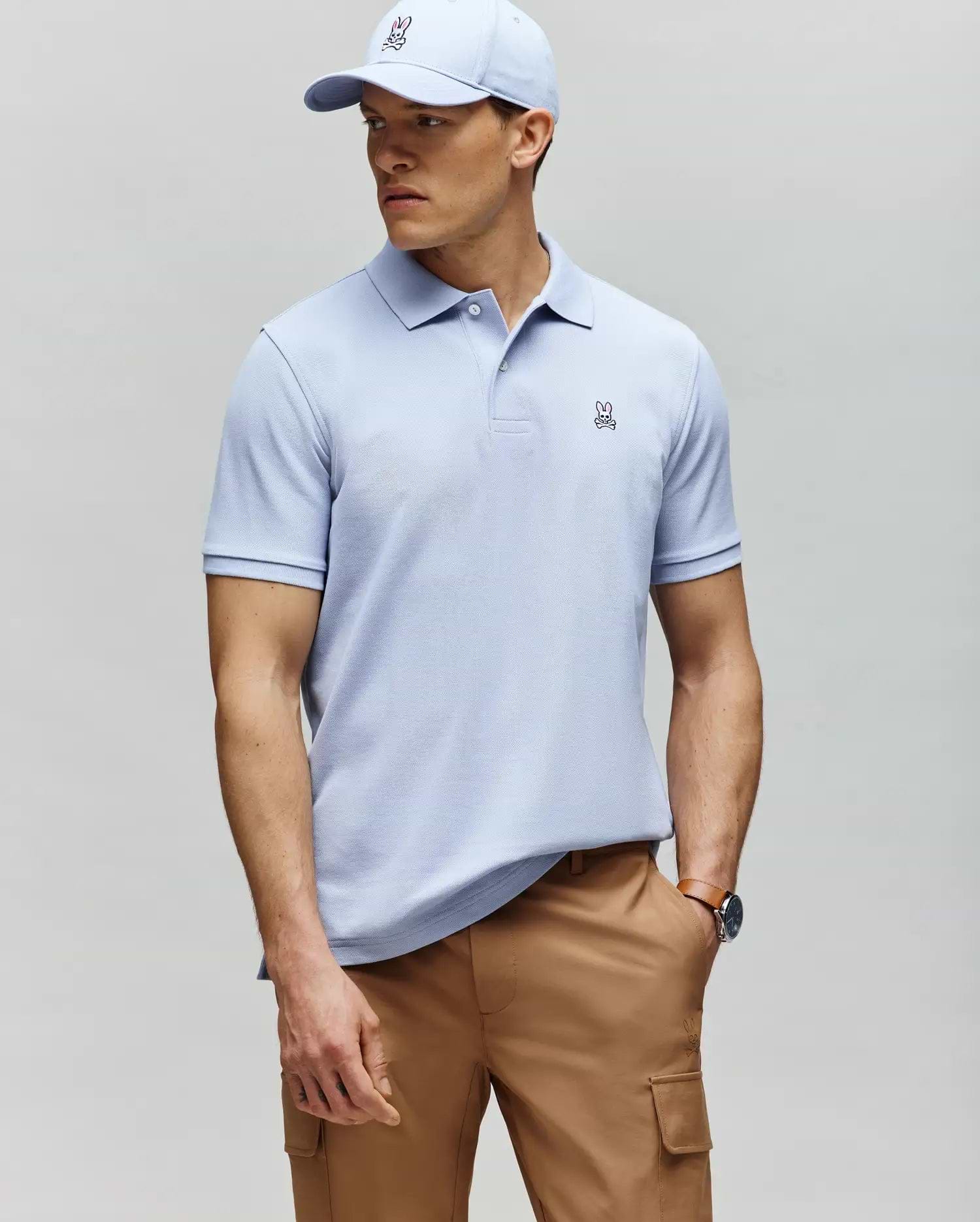 A man in a Psycho Bunny MENS CLASSIC PIQUE POLO - B6K001F200 in light blue with a small bunny logo, paired with a matching cap and tan cargo pants, stands against a gray background looking to the side.
