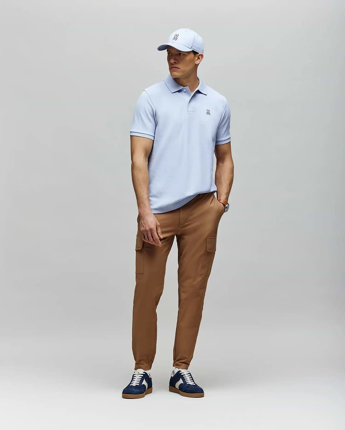 A man stands against a light gray background wearing a light blue Psycho Bunny MENS CLASSIC PIQUE POLO - B6K001F200, tan cargo pants, navy and white sneakers, and a matching light blue baseball cap.