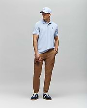 A man stands against a light gray background wearing a light blue Psycho Bunny MENS CLASSIC PIQUE POLO - B6K001F200, tan cargo pants, navy and white sneakers, and a matching light blue baseball cap.