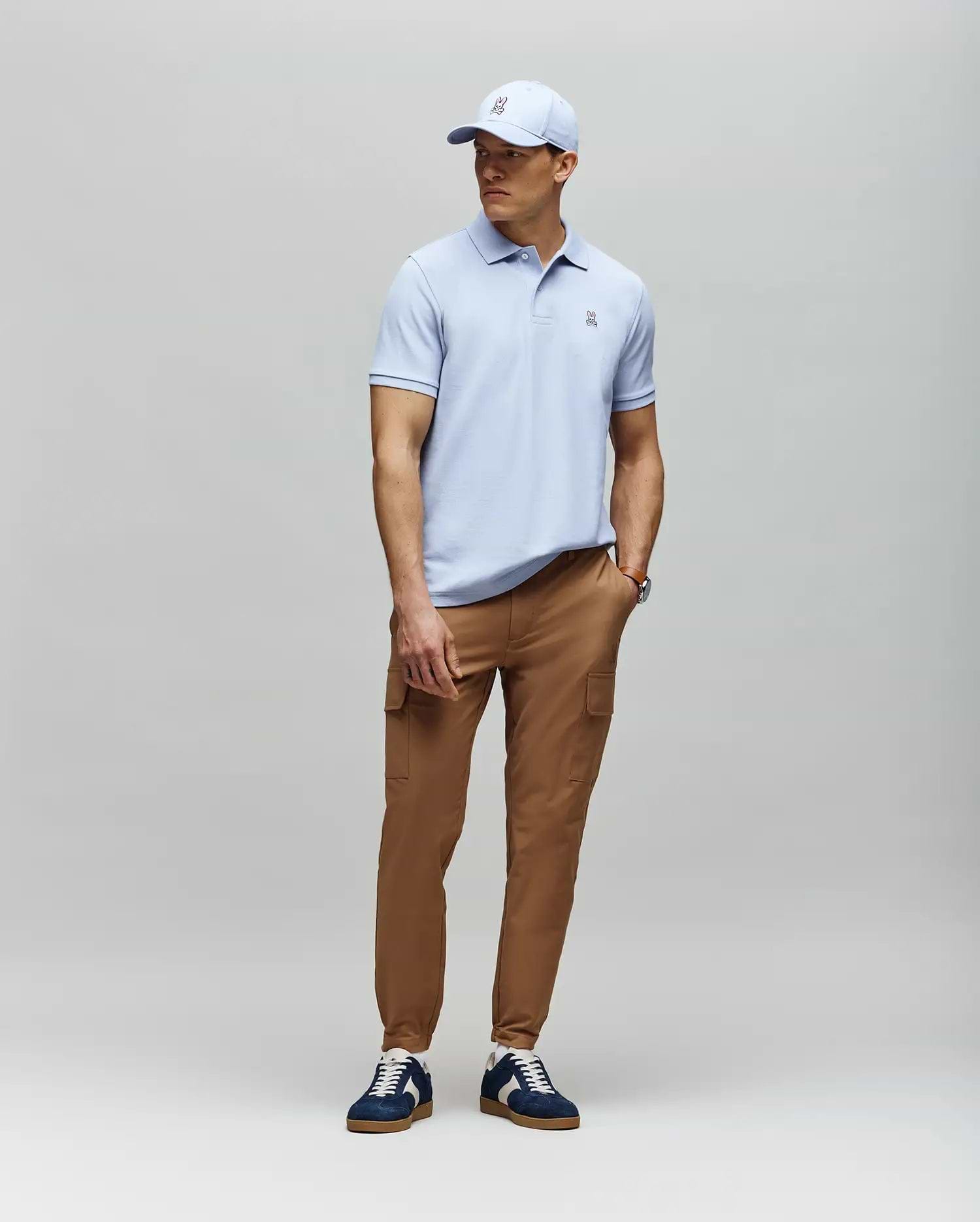 A man stands against a light gray background wearing a light blue Psycho Bunny MENS CLASSIC PIQUE POLO - B6K001F200, tan cargo pants, navy and white sneakers, and a matching light blue baseball cap.
