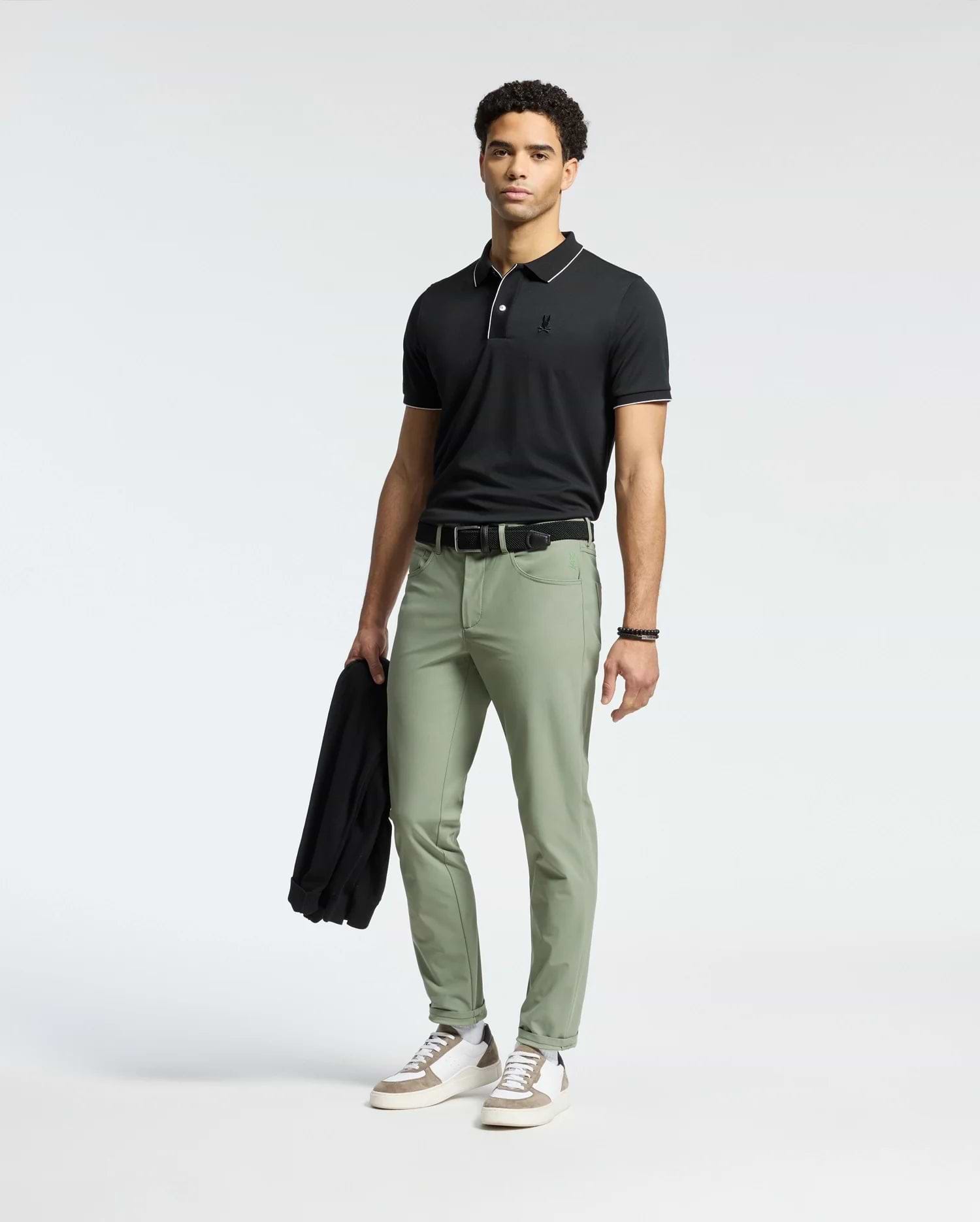 Dressed in a Psycho Bunny MENS BRYCE PERFORMANCE PIQUE POLO, black, with light green pants, the person holds a black jacket. They wear a bracelet on their left wrist and white sneakers with gray and brown accents, standing against a plain white background.