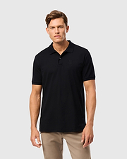 A man with short hair stands against a plain background, wearing the Psycho Bunny MENS JAMES PREMIUM PIQUE POLO SHIRT - B6K945D200 in black and beige pants. He faces the camera with a neutral expression.