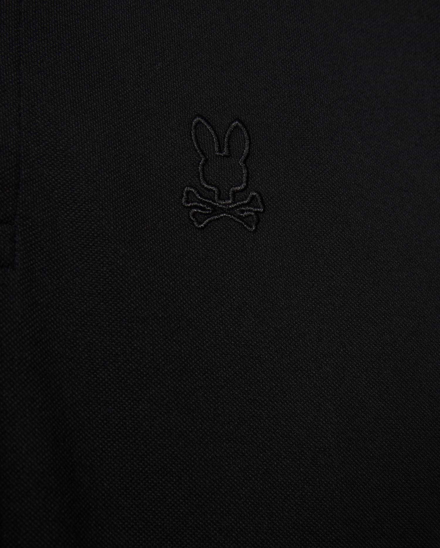 Close-up of the Psycho Bunny MENS JAMES PREMIUM PIQUE POLO SHIRT - B6K945D200 in black, showcasing an embroidered logo depicting a rabbit's head with crossbones underneath. The all-black logo blends subtly with the fabric, adding a refined touch. Made from premium Pima cotton, this durable shirt offers both style and comfort.