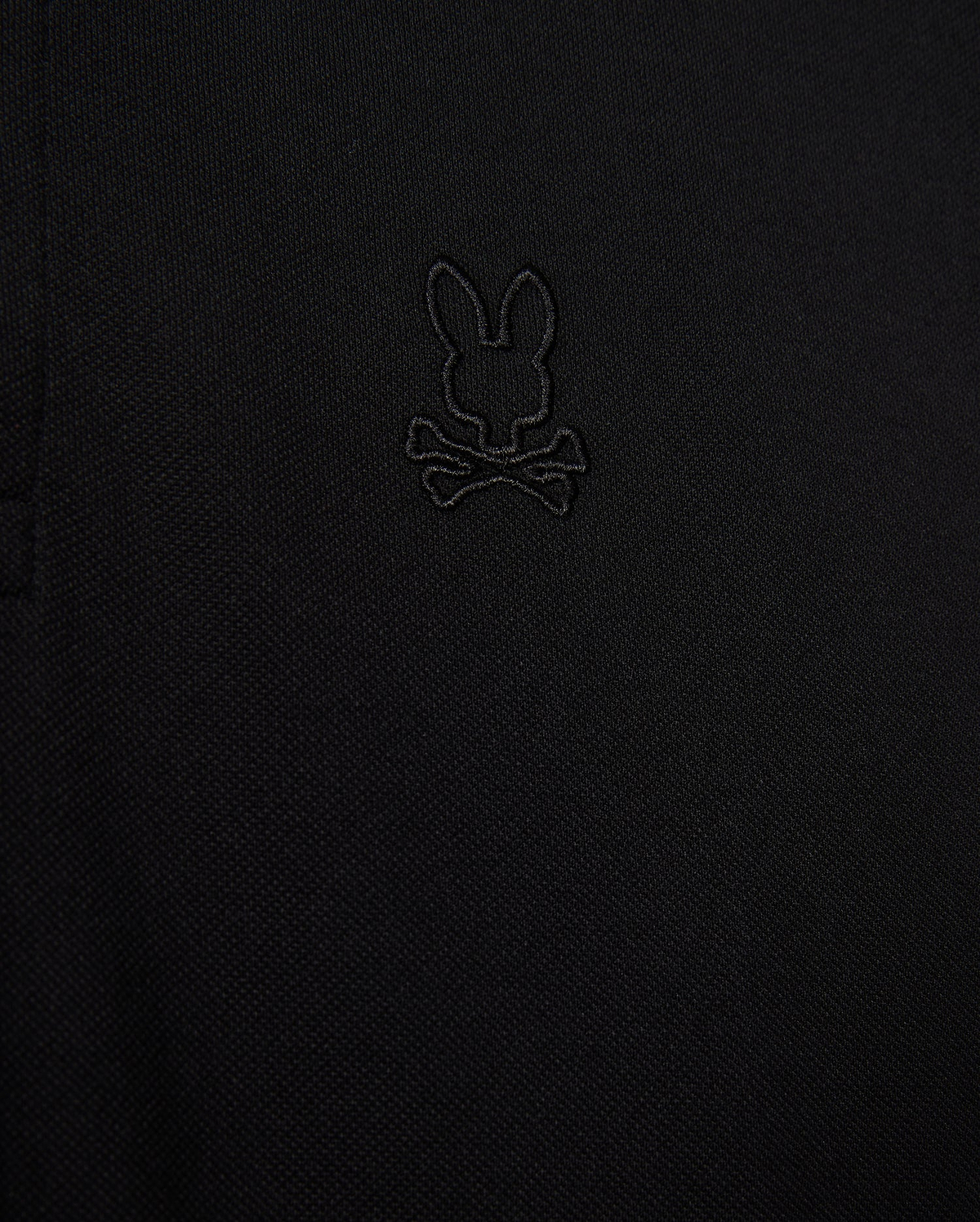 Close-up of the Psycho Bunny MENS JAMES PREMIUM PIQUE POLO SHIRT - B6K945D200 in black, showcasing an embroidered logo depicting a rabbit's head with crossbones underneath. The all-black logo blends subtly with the fabric, adding a refined touch. Made from premium Pima cotton, this durable shirt offers both style and comfort.