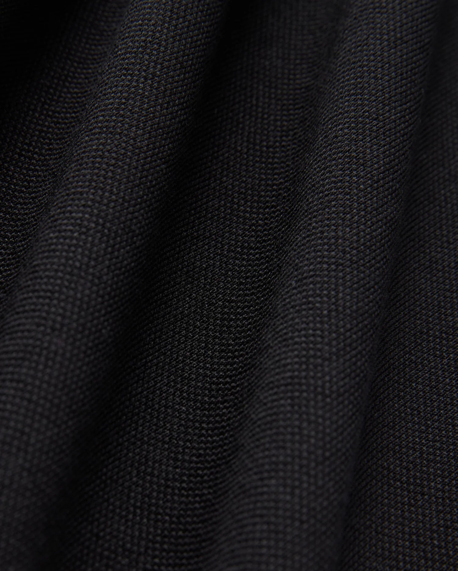 Close-up of the black Pima cotton fabric under soft lighting, showcasing the fine weave and subtle folds of Psycho Bunny's MENS JAMES PREMIUM PIQUE POLO SHIRT - B6K945D200. The surface appears smooth and uniform, with shadows emphasizing the gentle undulations typical of this high-quality polo shirt.