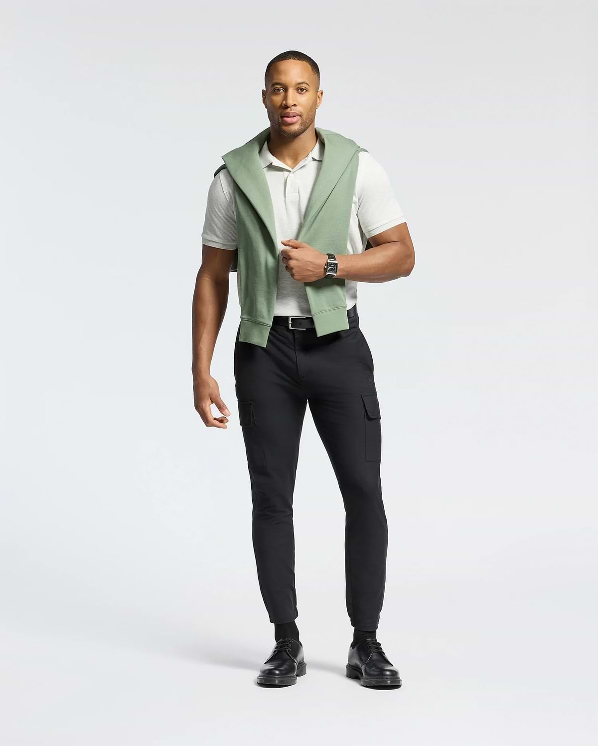 A man stands casually in a light gray Psycho Bunny MENS JAMES PIQUE POLO - B6K945D200, made of durable Pima cotton, paired with black pants and shoes. A green sweatshirt is draped over his shoulders, and he wears a wristwatch against a plain white background.