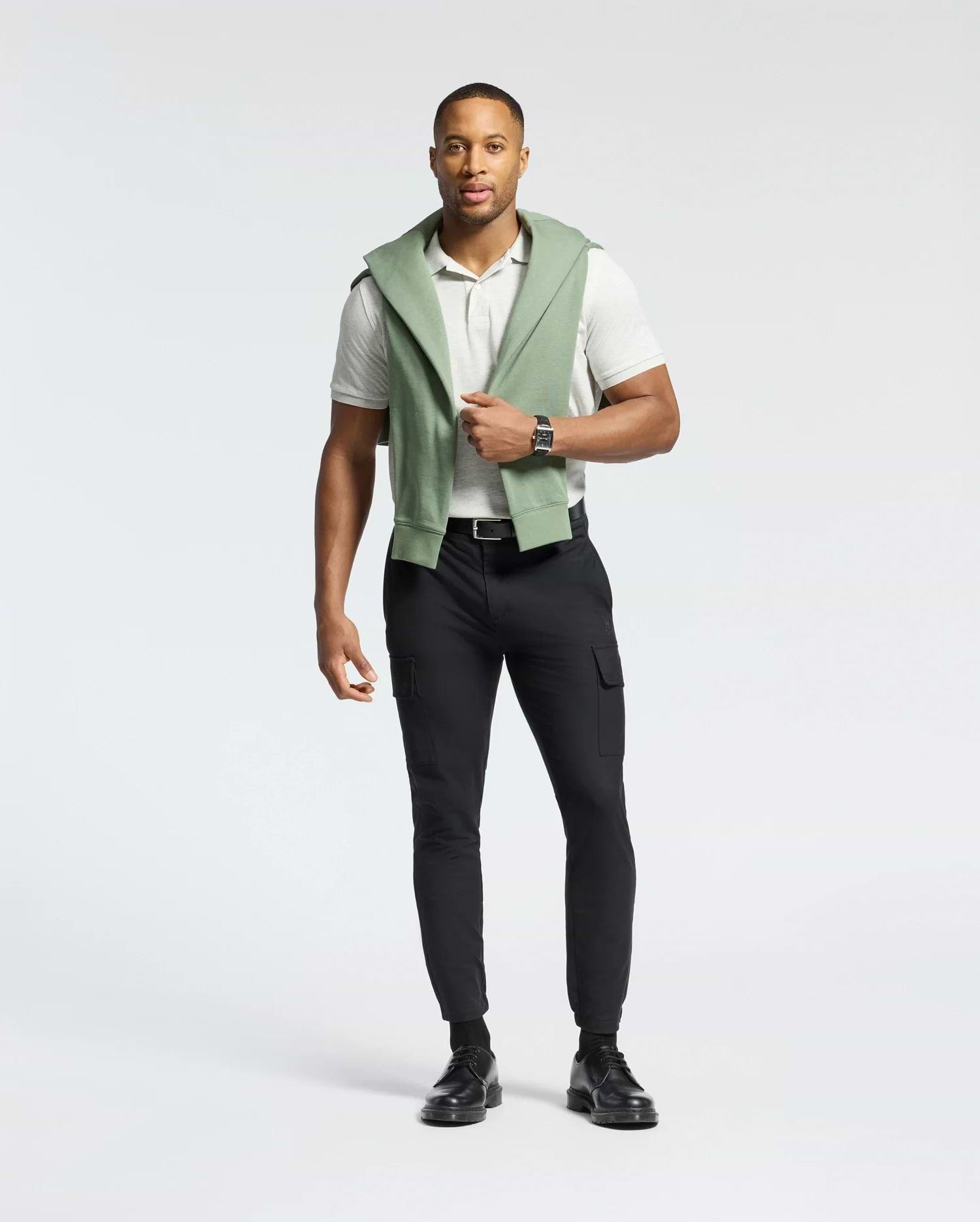 A man stands casually in a light gray Psycho Bunny MENS JAMES PIQUE POLO - B6K945D200, made of durable Pima cotton, paired with black pants and shoes. A green sweatshirt is draped over his shoulders, and he wears a wristwatch against a plain white background.