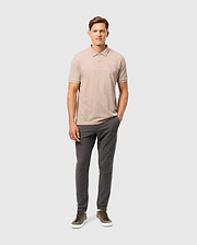 A man stands against a plain white background wearing a Psycho Bunny Men's James Premium Pique Polo Shirt (B6K945D200) made from Pima cotton, dark gray pants, and olive green sneakers. He has one hand in his pocket and is looking towards the camera with a neutral expression.