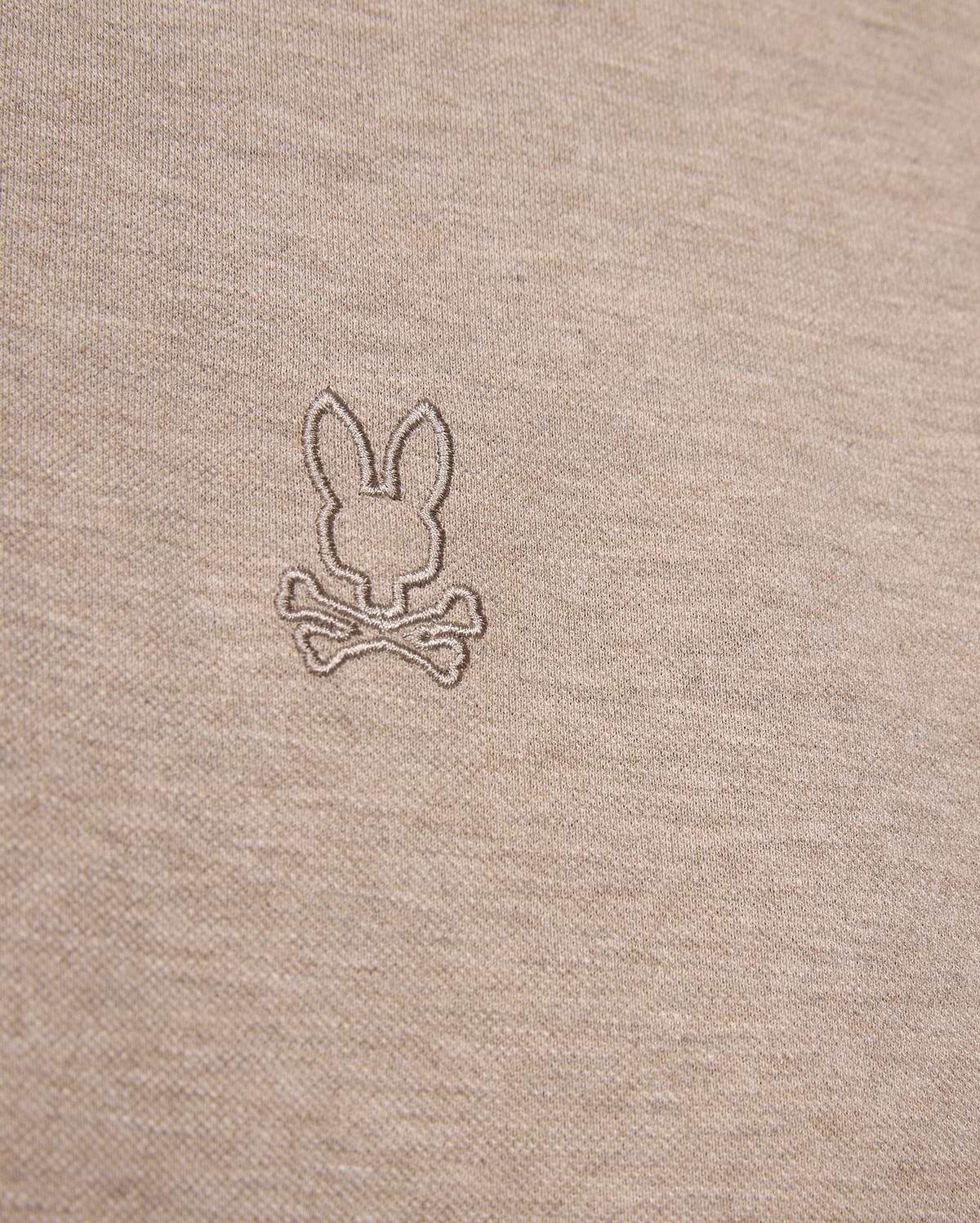 Close-up image of a beige fabric featuring an embroidered logo of a bunny head above two crossed bones, offering a playful twist on the classic skull and crossbones symbol. The embroidery, crafted in a slightly darker shade of beige, enhances the unique character of this durable Psycho Bunny James Premium Pique Polo Shirt - B6K945D200 made from premium Pima cotton.