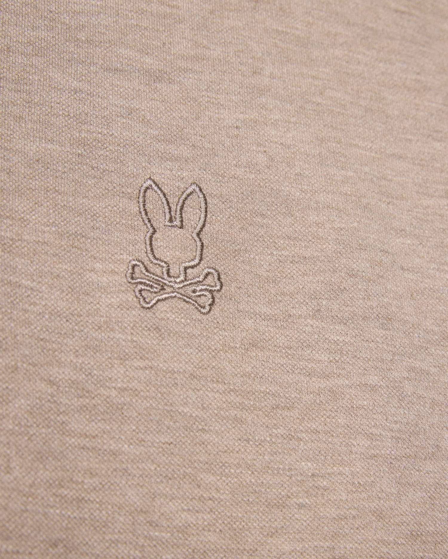 Close-up image of a beige fabric featuring an embroidered logo of a bunny head above two crossed bones, offering a playful twist on the classic skull and crossbones symbol. The embroidery, crafted in a slightly darker shade of beige, enhances the unique character of this durable Psycho Bunny James Premium Pique Polo Shirt - B6K945D200 made from premium Pima cotton.