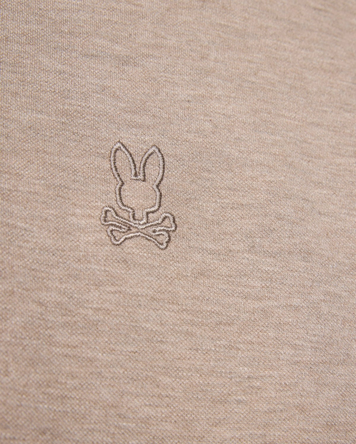 Close-up image of a beige fabric featuring an embroidered logo of a bunny head above two crossed bones, offering a playful twist on the classic skull and crossbones symbol. The embroidery, crafted in a slightly darker shade of beige, enhances the unique character of this durable Psycho Bunny James Premium Pique Polo Shirt - B6K945D200 made from premium Pima cotton.