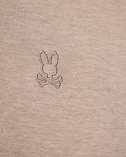Close-up image of a beige fabric featuring an embroidered logo of a bunny head above two crossed bones, offering a playful twist on the classic skull and crossbones symbol. The embroidery, crafted in a slightly darker shade of beige, enhances the unique character of this durable Psycho Bunny James Premium Pique Polo Shirt - B6K945D200 made from premium Pima cotton.