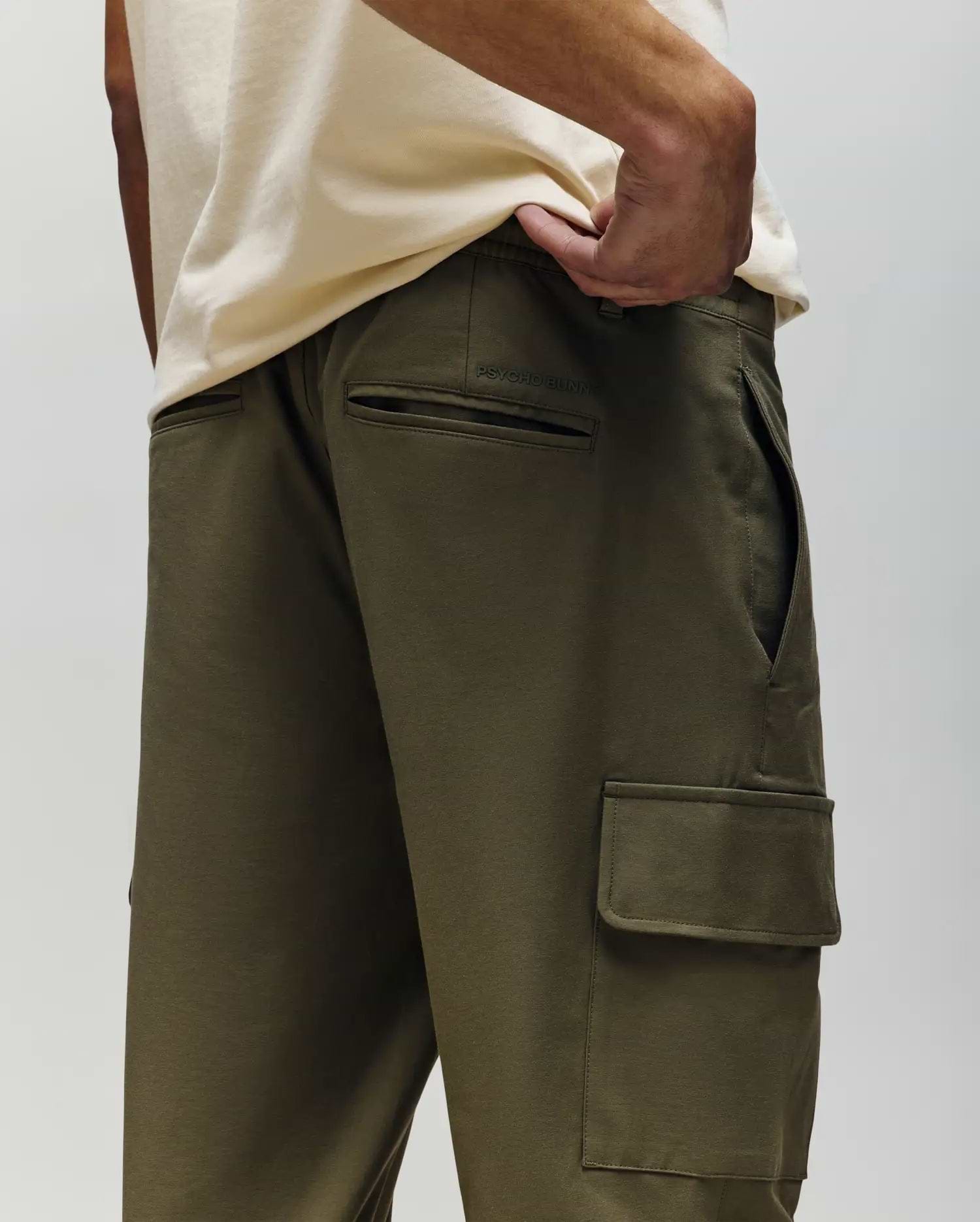 A person models the Psycho Bunny MENS LLOYD CARGO JOGGER - B6P159B200 in olive green, paired with a cream shirt, highlighting the jogger’s side and back pockets, relaxed fit, and detachable keychain on a belt loop.