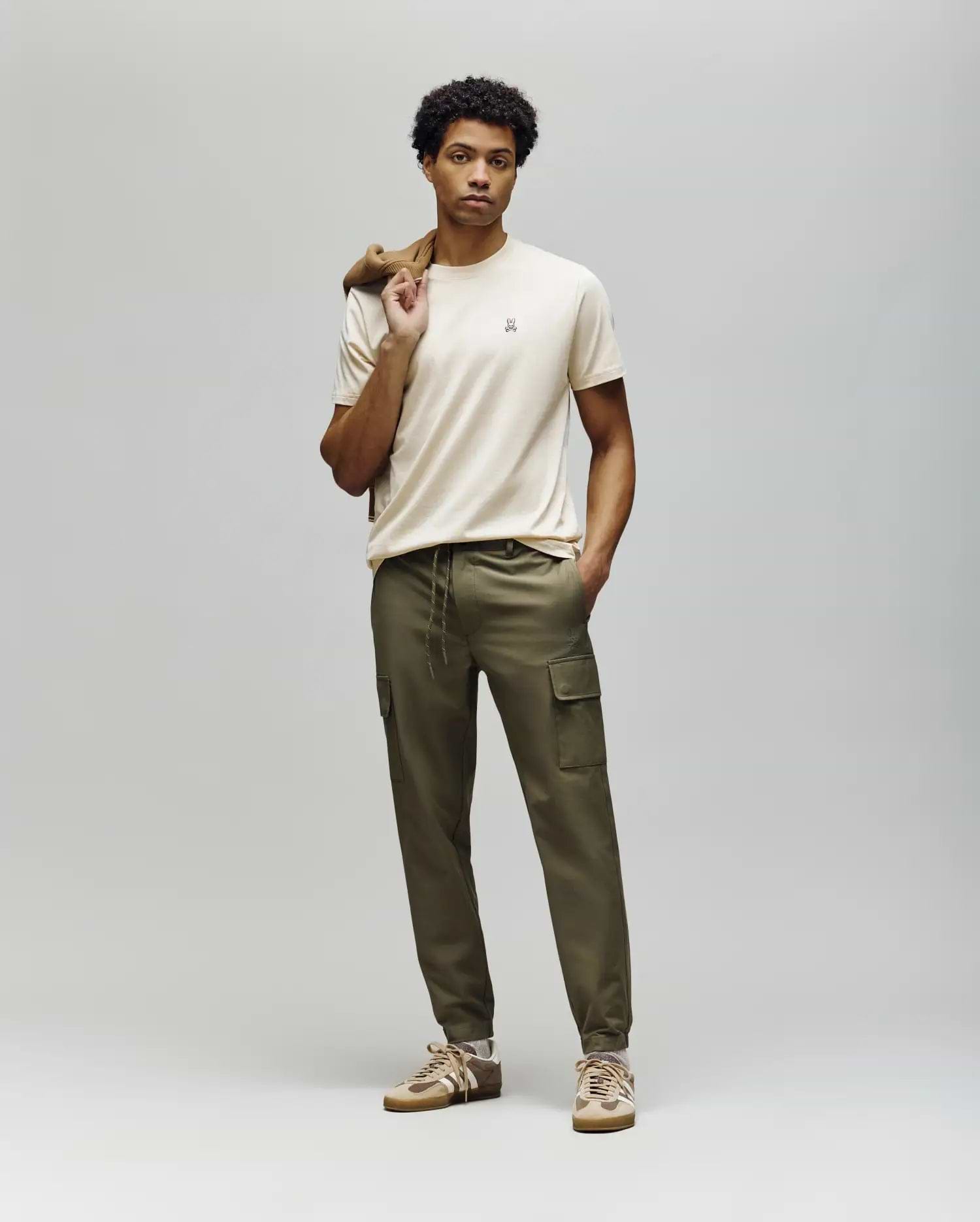 A man stands against a plain background wearing Psycho Bunny's MENS LLOYD CARGO JOGGER - B6P159B200, a beige t-shirt, tan sneakers, and holds a brown jacket over his shoulder, gazing straight ahead with a neutral expression.