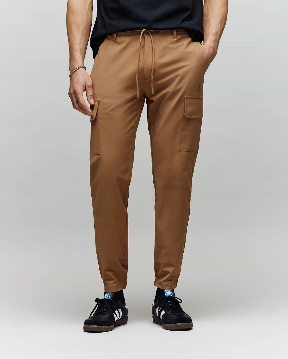 A person wearing Psycho Bunny MENS LLOYD CARGO JOGGER - B6P159B200 with side pockets and subtle embroidery, paired with a black t-shirt and black Adidas sneakers with white stripes, stands against a plain gray background.