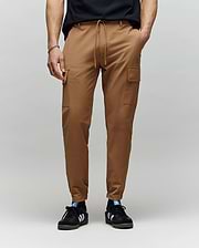 A person wearing Psycho Bunny MENS LLOYD CARGO JOGGER - B6P159B200 with side pockets and subtle embroidery, paired with a black t-shirt and black Adidas sneakers with white stripes, stands against a plain gray background.