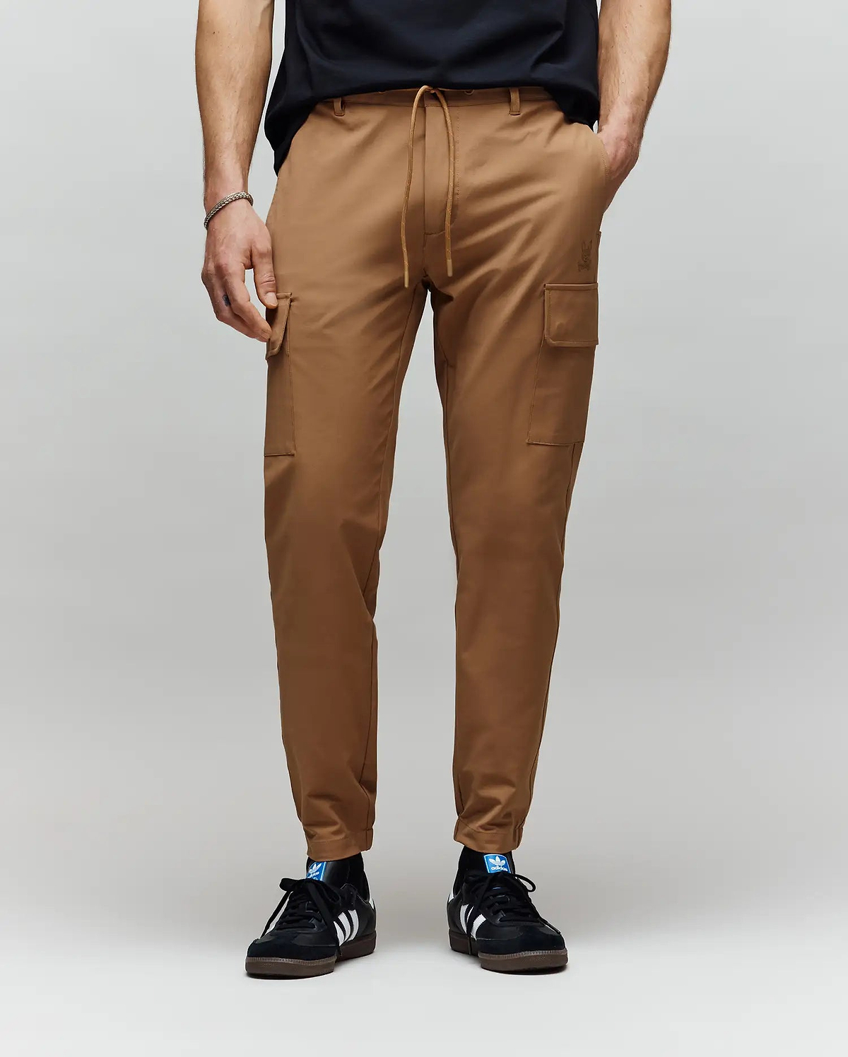 A person wearing Psycho Bunny MENS LLOYD CARGO JOGGER - B6P159B200 with side pockets and subtle embroidery, paired with a black t-shirt and black Adidas sneakers with white stripes, stands against a plain gray background.